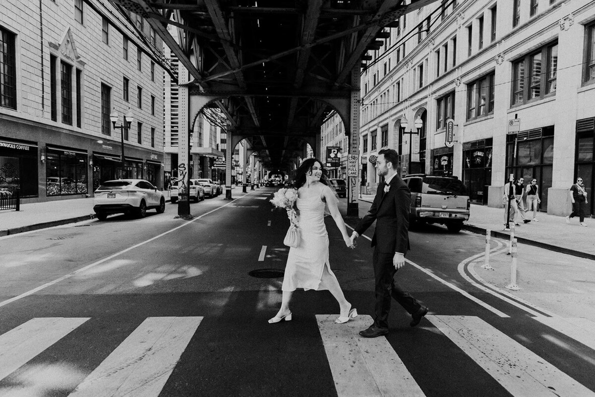 Just Married photo session couple walks under Chicago L as they cross street. They hold hands and she is looking back at him.