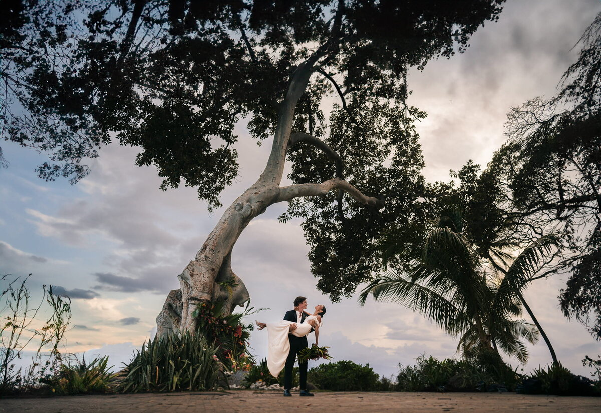 In a whimsical moment, the groom lifts the bride, surrounded by the grandeur of a big tree and a dramatic sky, adding magic to their special day.