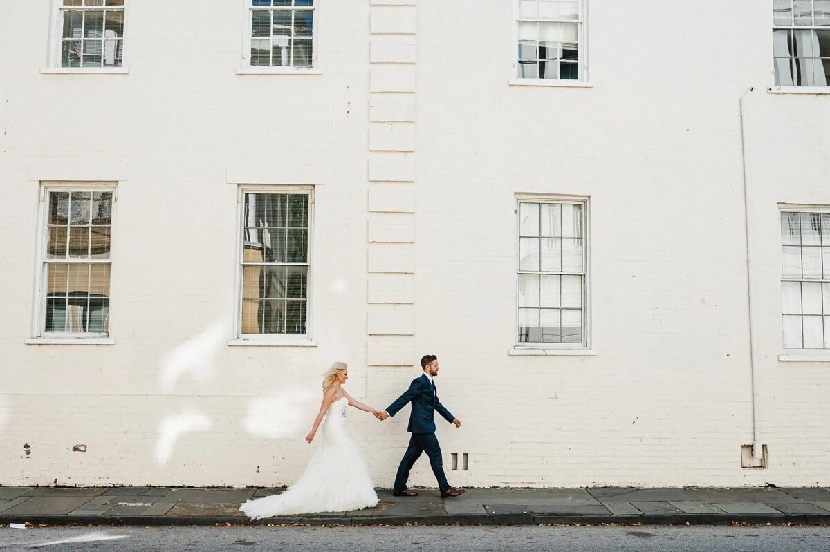 A bride and groom holding hands and walking along a sidewalk.