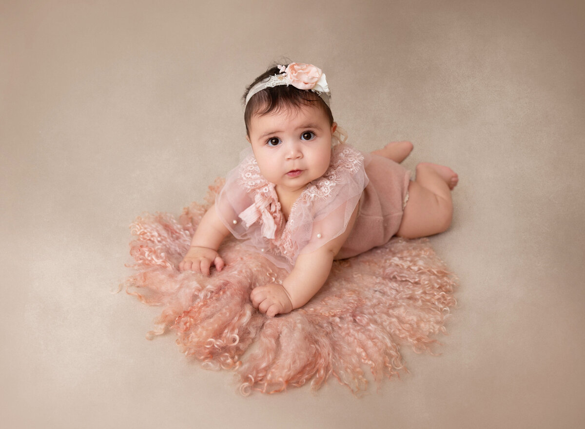 Baby girl is lying on her belly for a 3-month baby milestone photoshoot. She is wearing a pink knit romper with organza and pearl detail and is looking up at the camera.