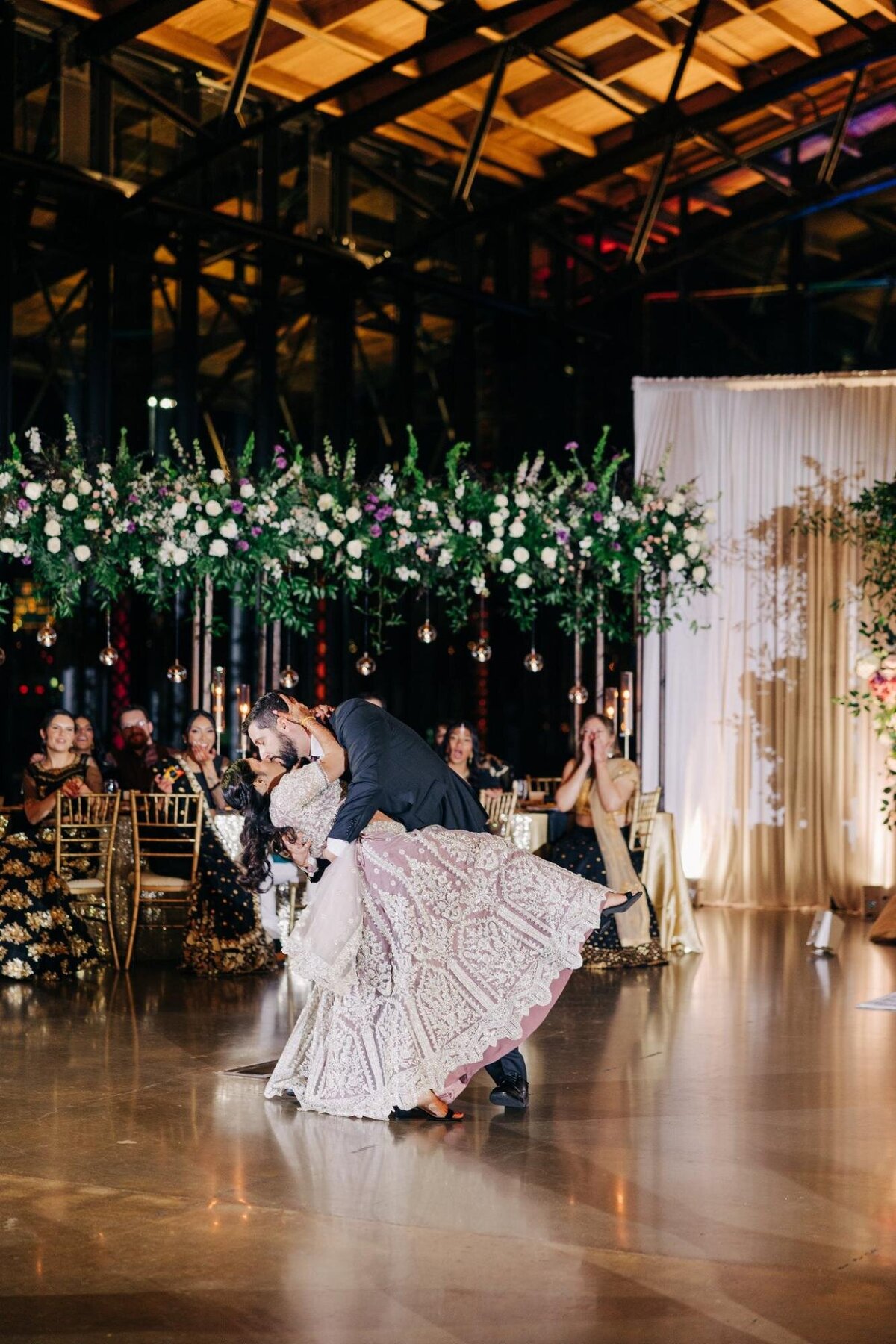 A groom dips the bride during their first dance at a wedding reception, surrounded by floral decorations and guests.