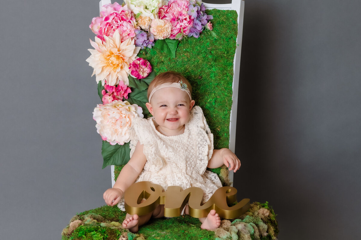 Cake Smash Photographer, a baby sits on a flowery chair and smiles