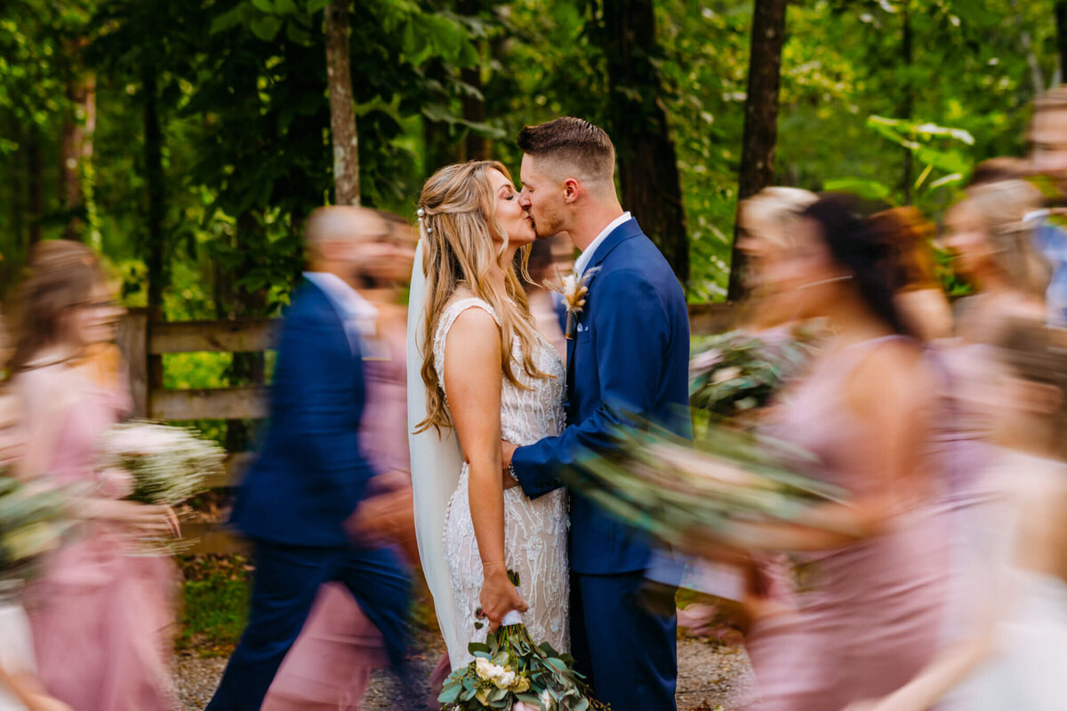 Photo of a bride and groom kissing while the wedding party walks around them in a blur