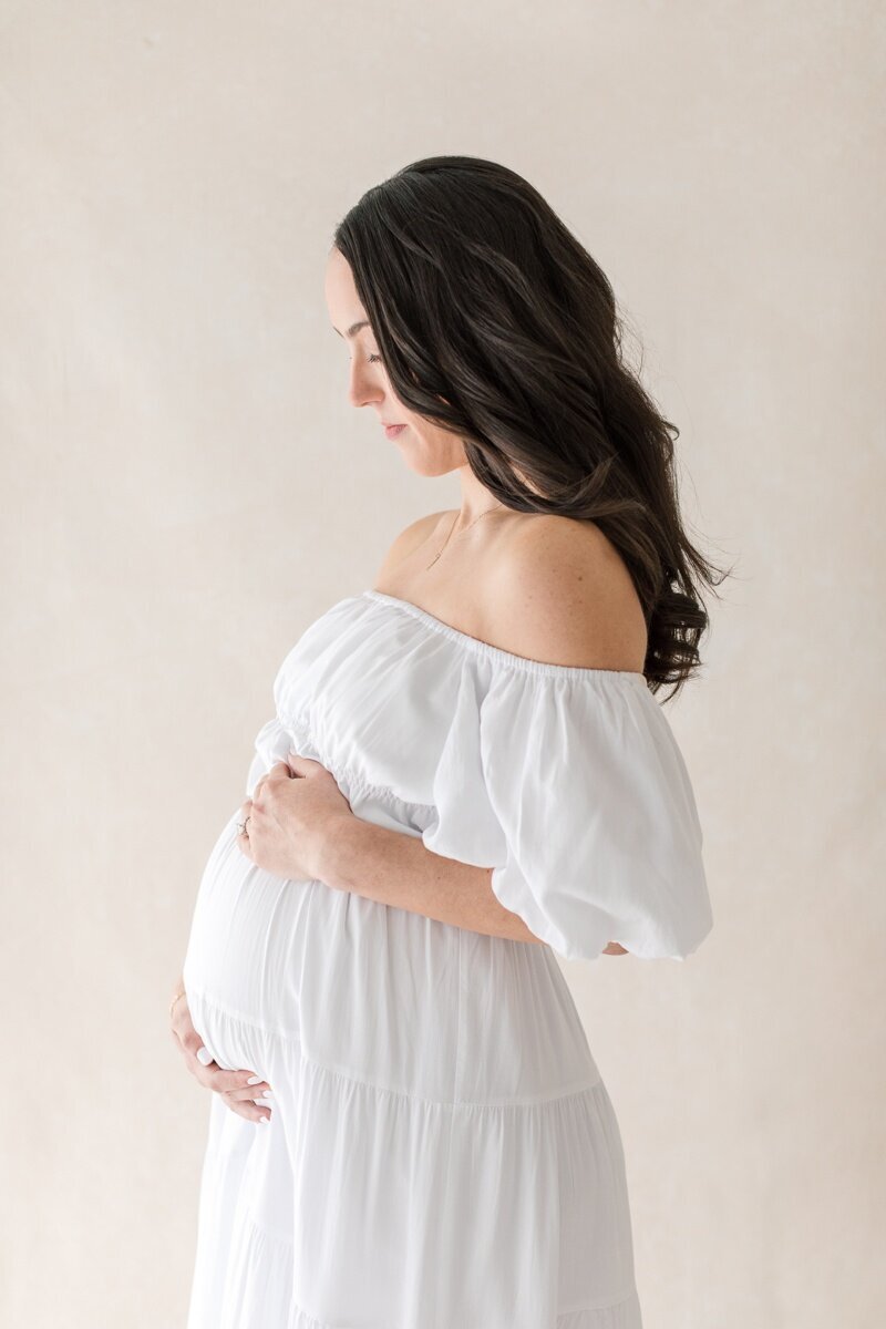 The profile view of a pregnant woman wearing a white off the shoulder dress holds her stomach as she gazes down at it during her Lawrenceville NJ Maternity Photography session