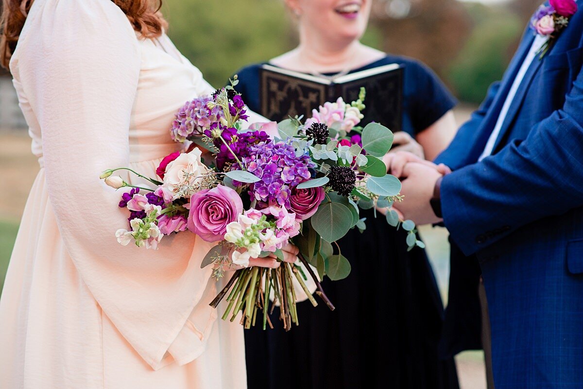 Detail photo of the bridal bouquet. The bouquet has pink, purple ivory, blush and burgundy flowers. The bride is wearing a boho ivory dress with bell sleeves and the groom is wearing a cobalt blue suit with a hot pink and blush boutonniere. The officiant stands behind them  in a back dress.