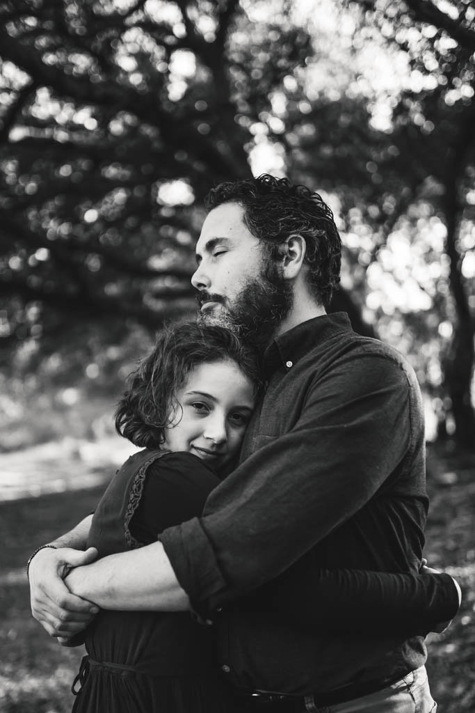 Lifestyle portrait of father embraces daughter with his eyes closed and girl looking at camera