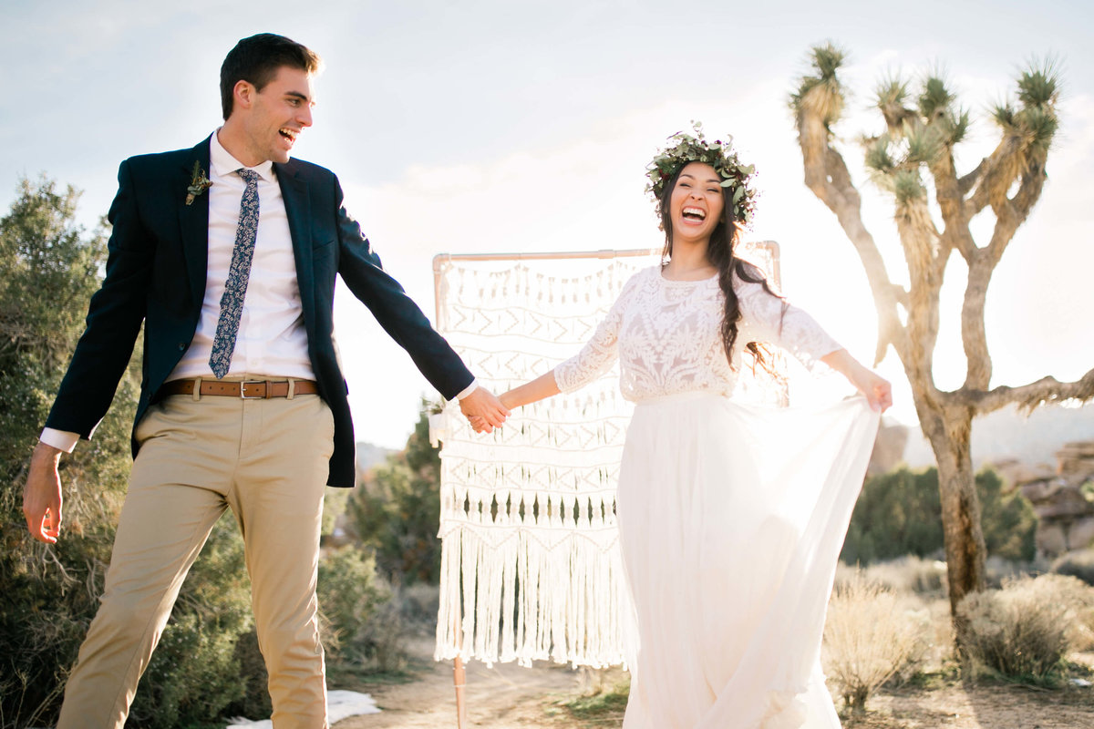 the couple laughs in front of their ceremony backdrop and joshua trees. the bride holds the corner of her skirt in the air