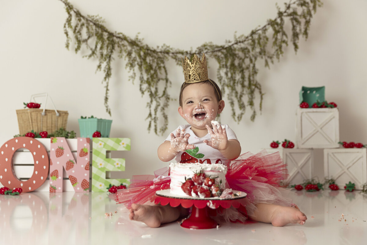 A young toddler girl in a red onesie tutu happily laughs while smashing a cake in a studio