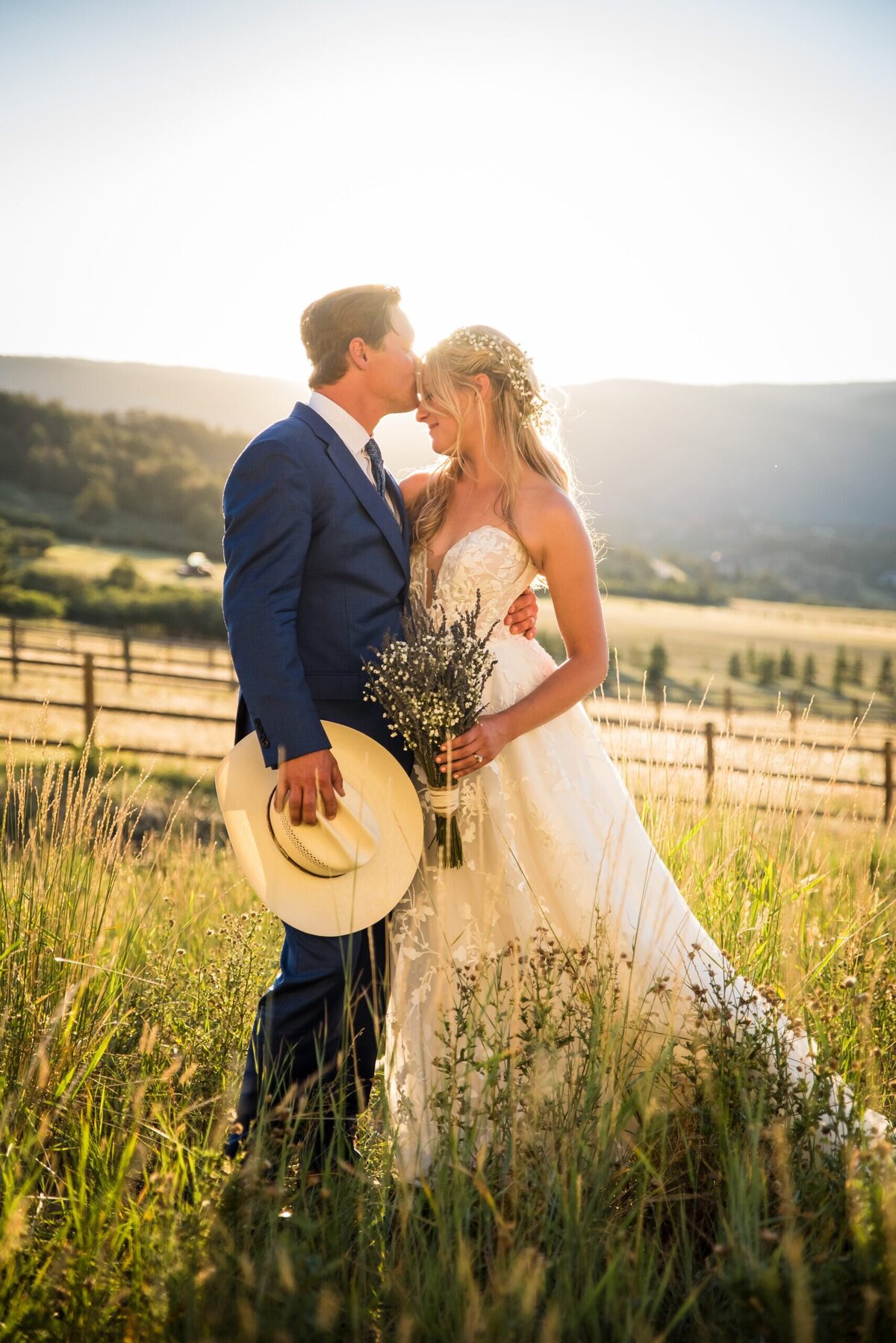 A groom holds his cowboy hat as he kisses his bride's forehead at golden hour.