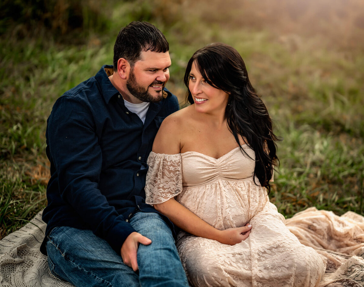 An adorable expecting couple sit on a blanket in a field of grass
