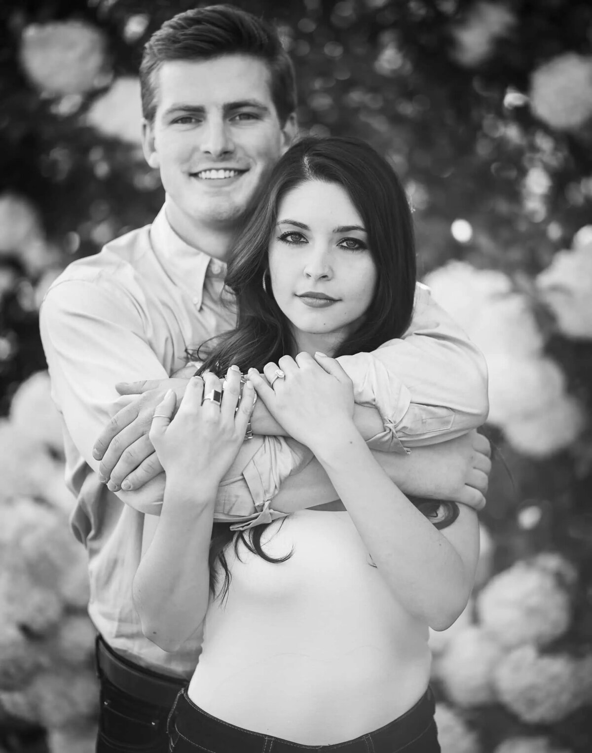 Black and white image of a smiling couple embracing, with the woman's hands on the man's chest and a backdrop of blooming flowers