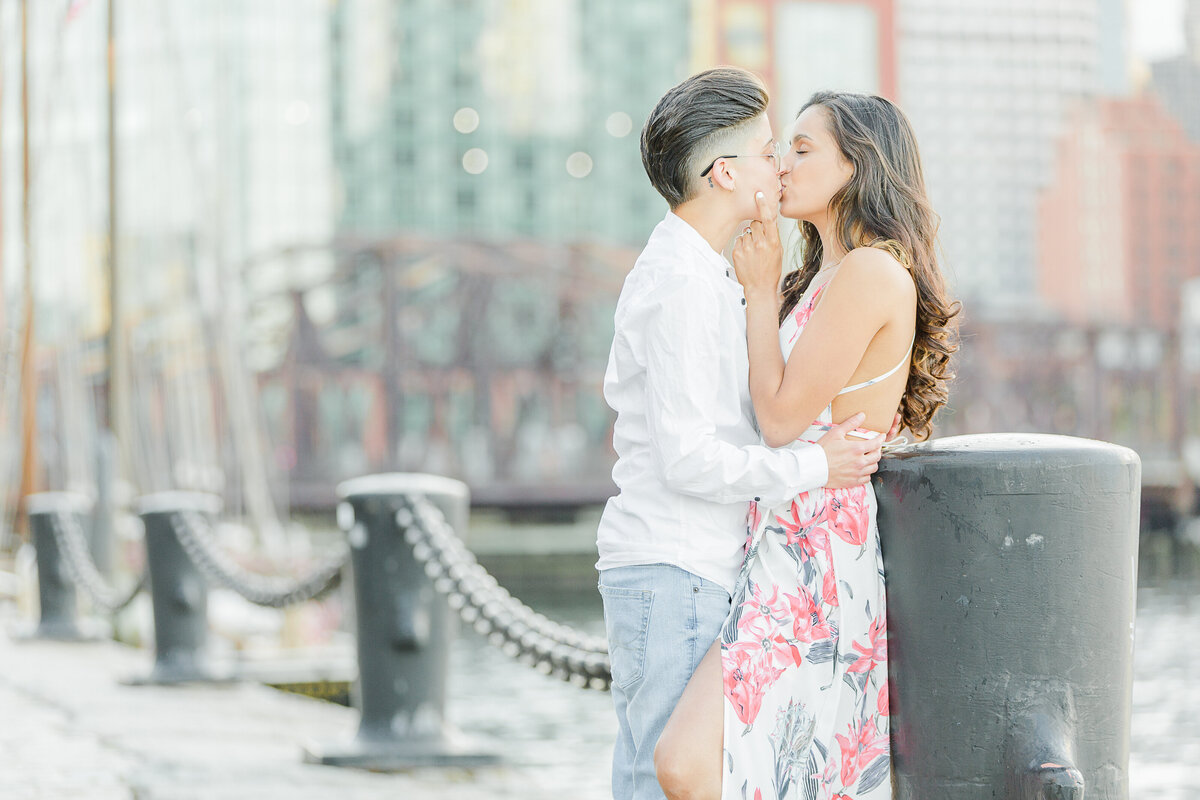 A couple shares a kiss at the Boston Seaport for their engagement photos. The woman is leaning against a large pillar. Captured by best MA engagement photographer Lia Rose Weddings