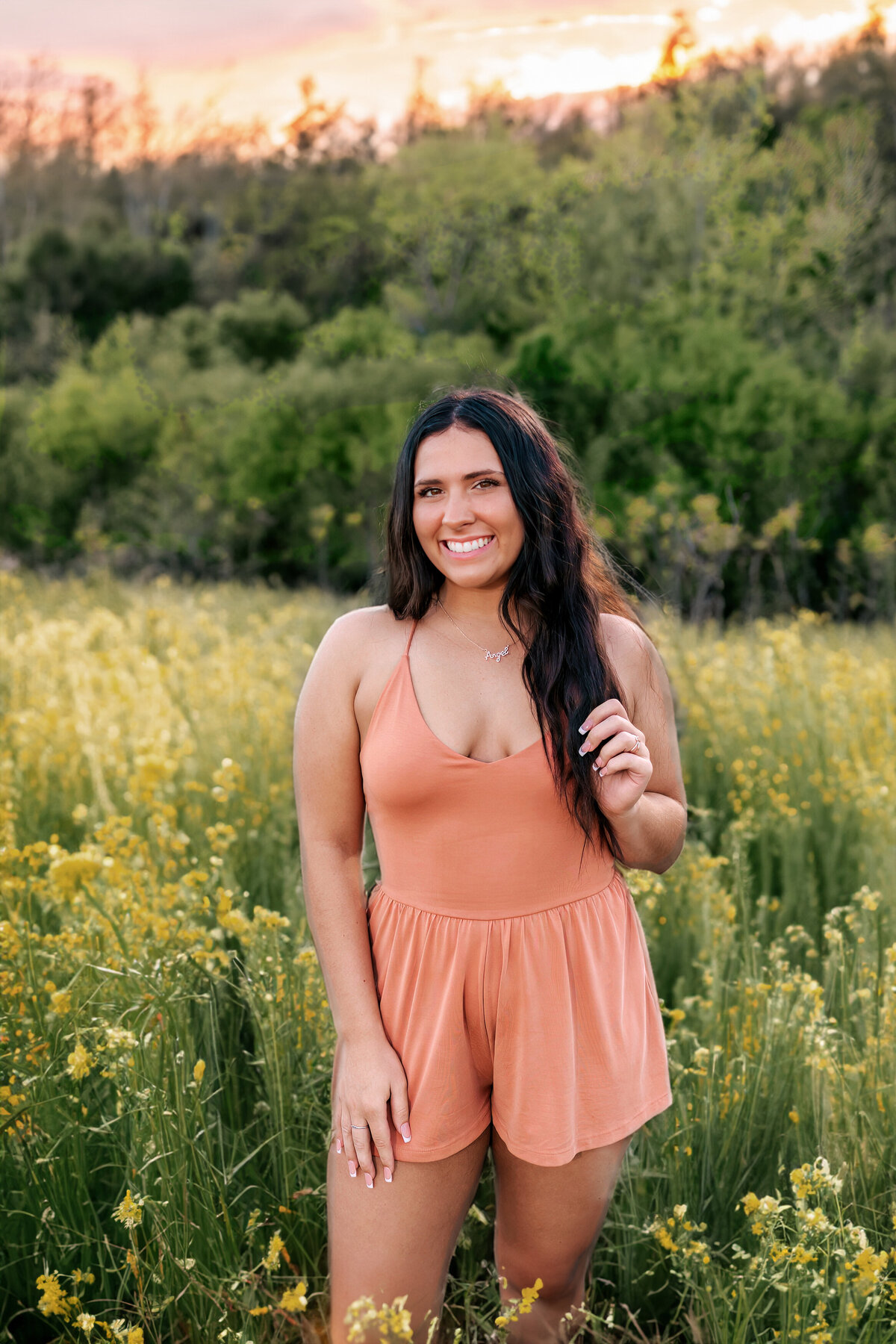 A beautiful girl is posing for her senior portraits in a field of wild flowers and tall grass at sunset.
