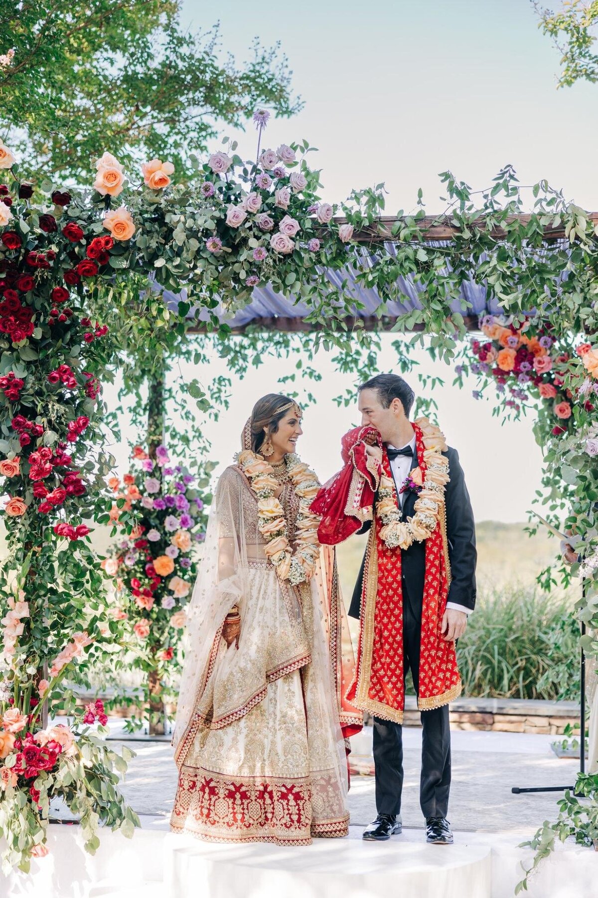 A bride and groom in traditional indian wedding attire exchange smiles under a floral archway outdoors.