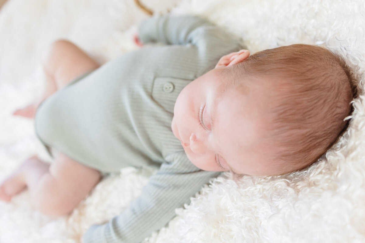 A newborn baby boy in a green onesie sleeping on a fluffy blanket during his NOVA newborn photos.