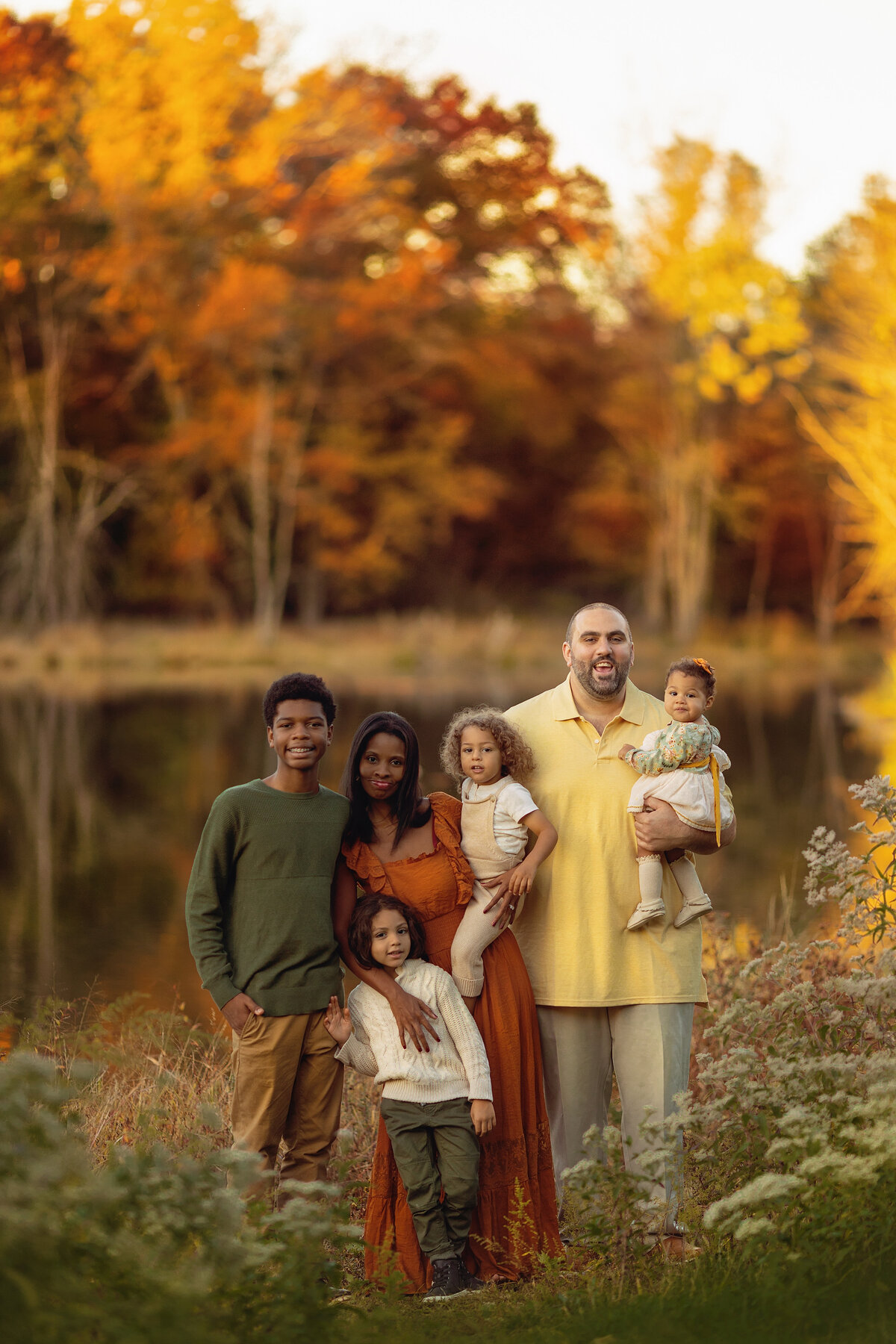 A mom and dad stand by a lake in a park in fall at sunset with their three toddlers and teenage son