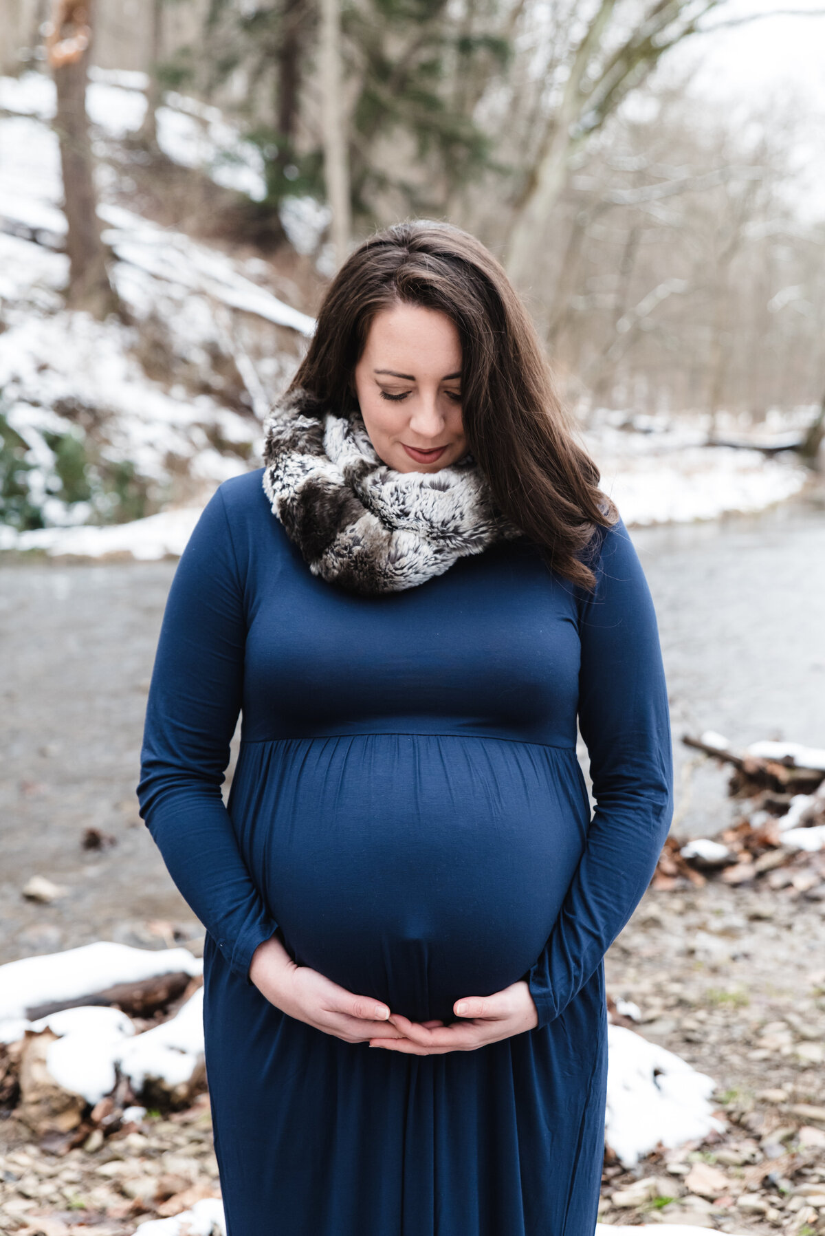 pregnant mom in blue dress hold belly in front of creek during winter
