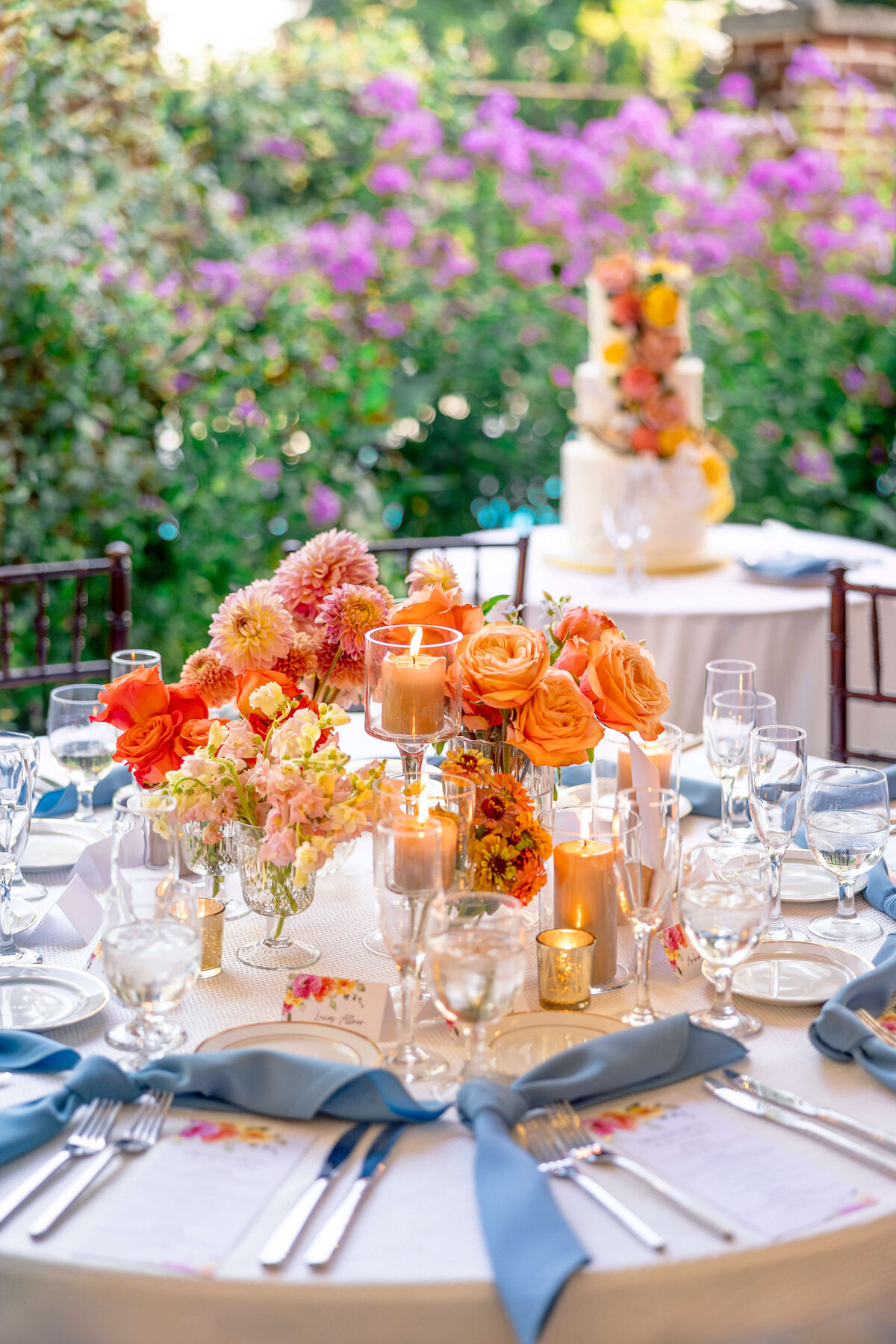 Elegant outdoor wedding table setting with orange and pink flowers, candles, and blue napkins. A white tiered cake adorned with flowers is in the background, surrounded by greenery and purple blooms.