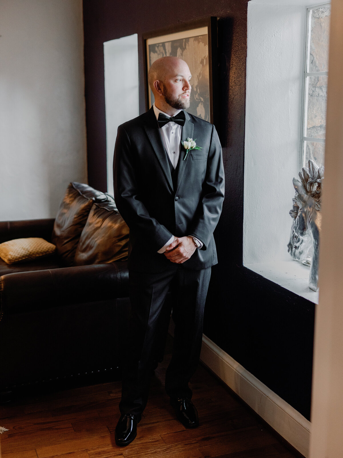 A groom stands near a window in his wedding suit with his hands held in front of his body