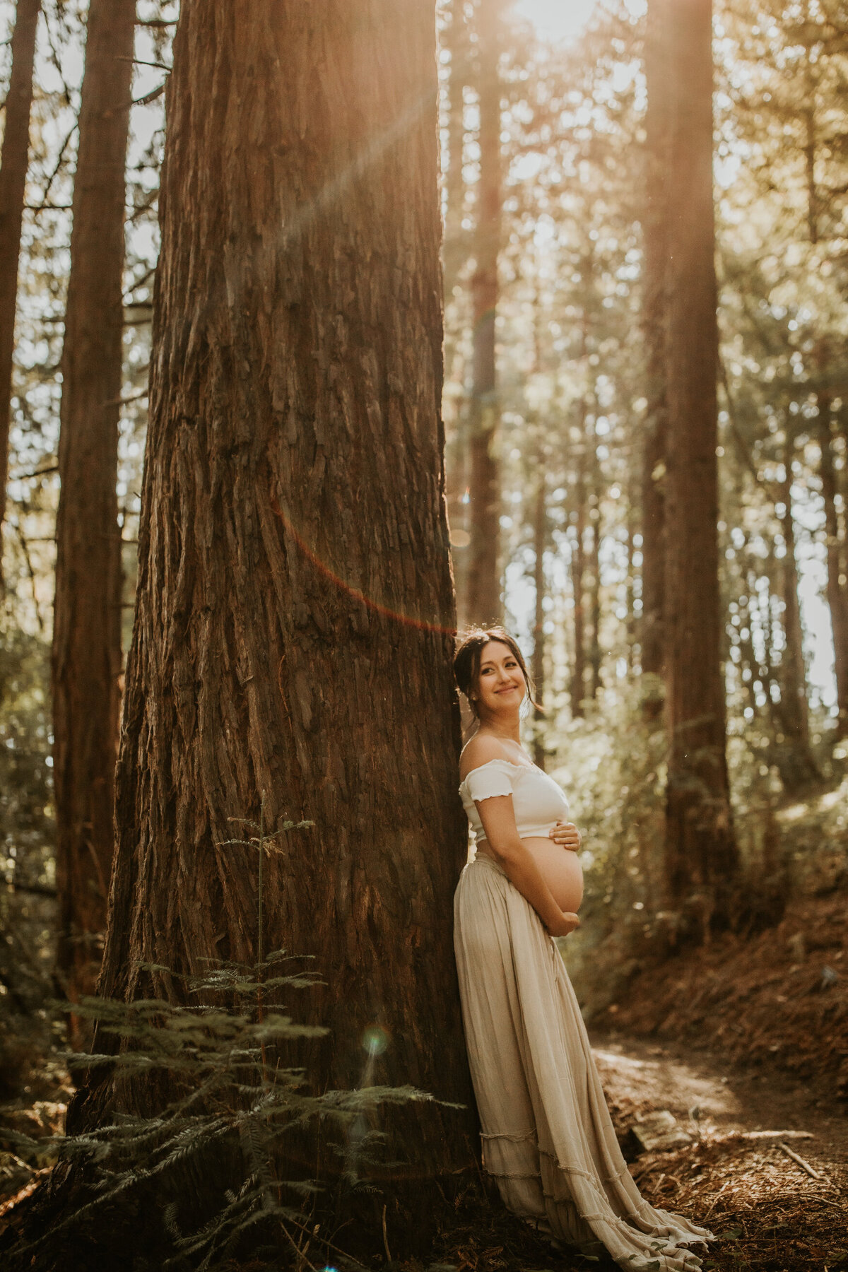 Bay Area maternity photoshoot in East Bay redwoods with mom leaning against tree and holding her bare belly while smiling at camera