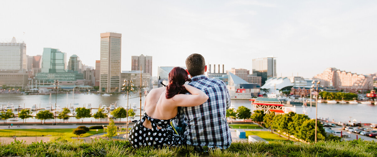 A couple sitting in the grass at a park overlooking a city skyline.
