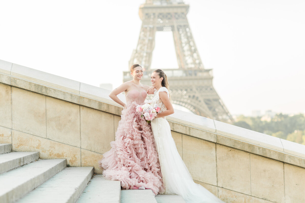 Bride and maid of honour stand on stone steps in Paris, France with the Eiffel Tower directly behind them. They are laughing together. Captured by destination wedding photographer Lia Rose Weddings