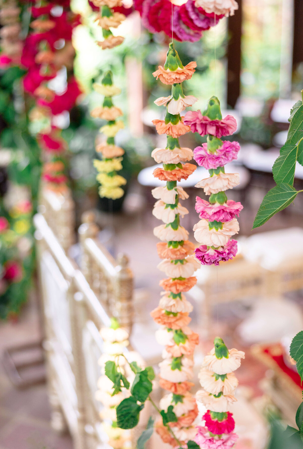 Colorful flower garlands hang vertically, with shades of pink, peach, yellow, and white. Green leaves accompany the garlands, creating a vibrant and festive atmosphere. A blurred background adds a sense of depth to the image.