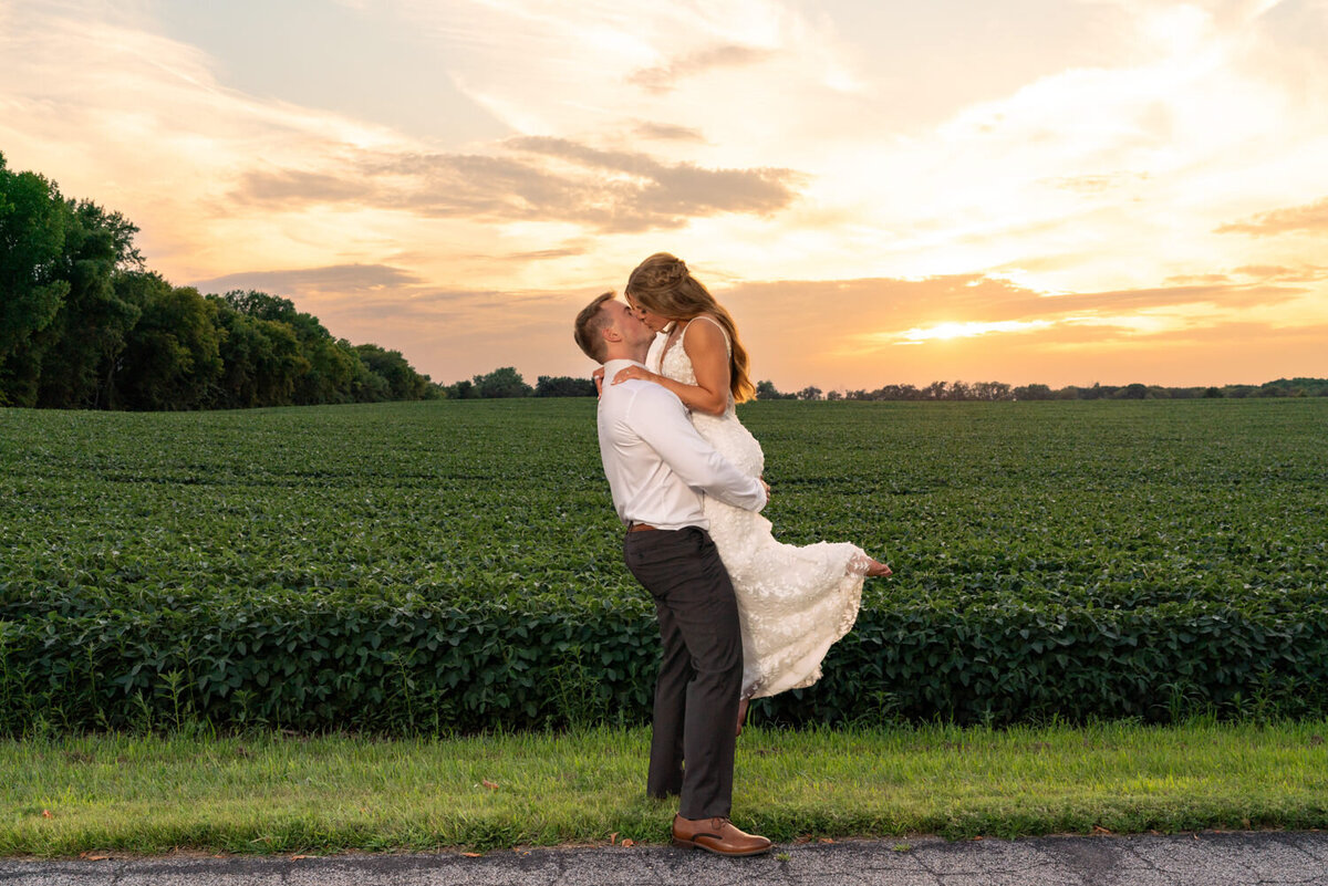 Groom lifts and kisses bride at sunset in front of a field in Minnesota.