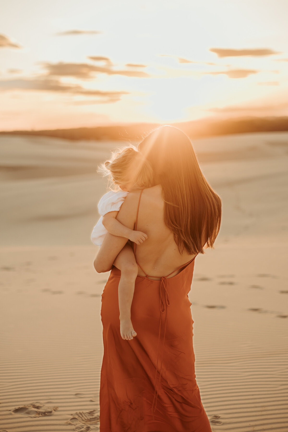 Little girl has her blonde head buried into her mum's shoulder while mum gazes towards the sun so her back is shown to the camera.