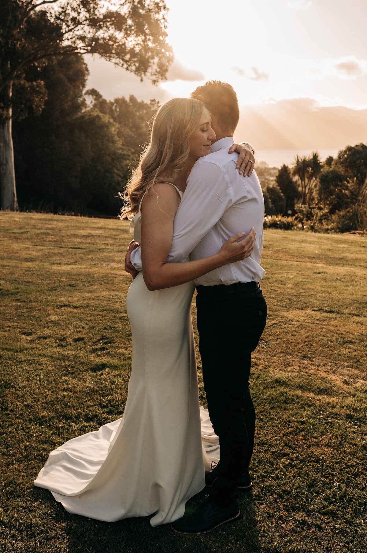 bride and groom hug at golden sunset on the lawn at mt vernon lodge overlooking akaroa harbour