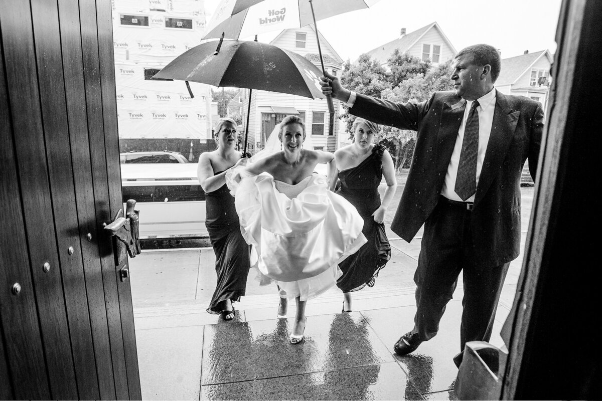 A bride huddles underneath an umbrella while it rains on her wedding