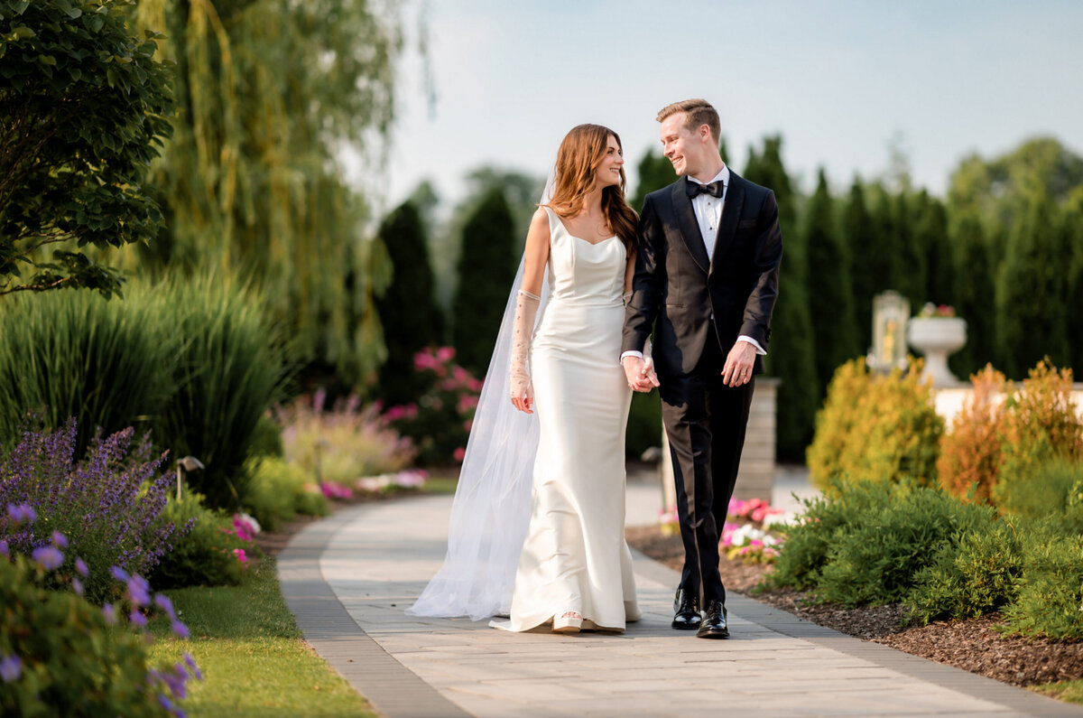 Bride and groom holding hands and walking down a walkway in the gardens of The Mansion on Main Street