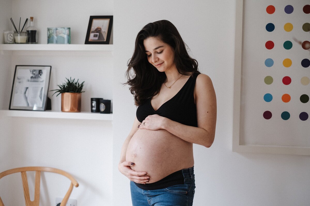 Pregnant woman wearing black bra and jeans stands in her dining room and holds her baby bump