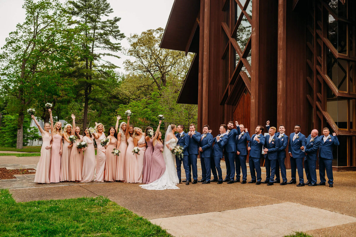 photo of a bride and groom kissing while the bridesmaids and groomsmen cheer in front of a wooden building