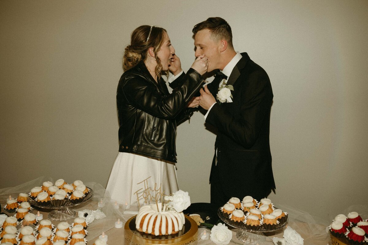 Bride in a leather jacket feeds groom a cupcake at a wedding reception table filled with various desserts, coordinated by a top wedding planner in Des Moines.