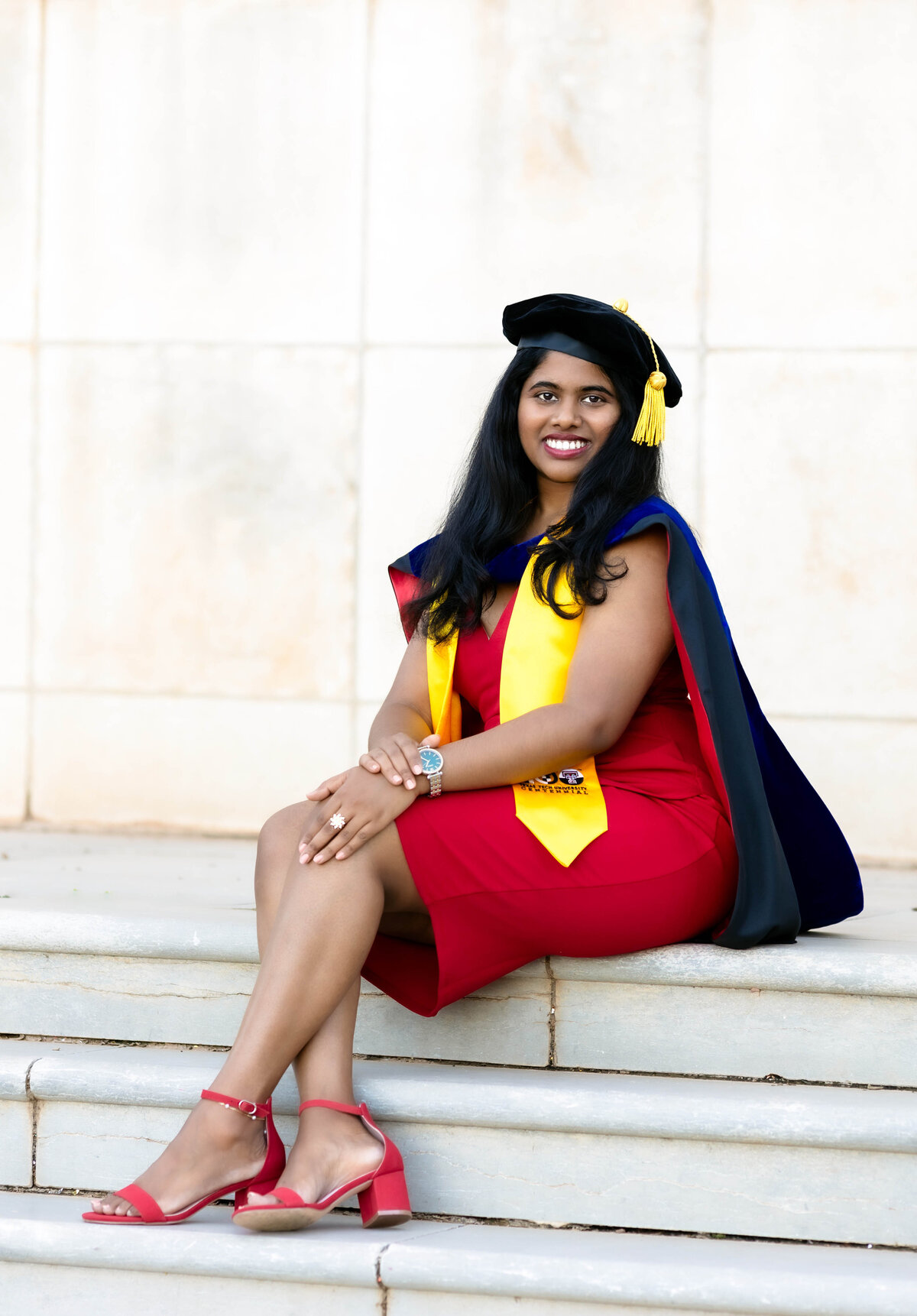 Indian college graduate session with woman wearing  a red dress and red shoes with a yellow stole seated while smiling at the camera