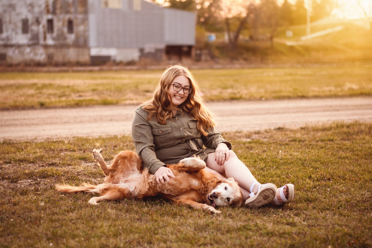A teenager in an olive dress sits in the grass and plays with a dog as the sunset illuminates her golden hair, captured by a fine art senior photographer in Bemidji, Minnesota