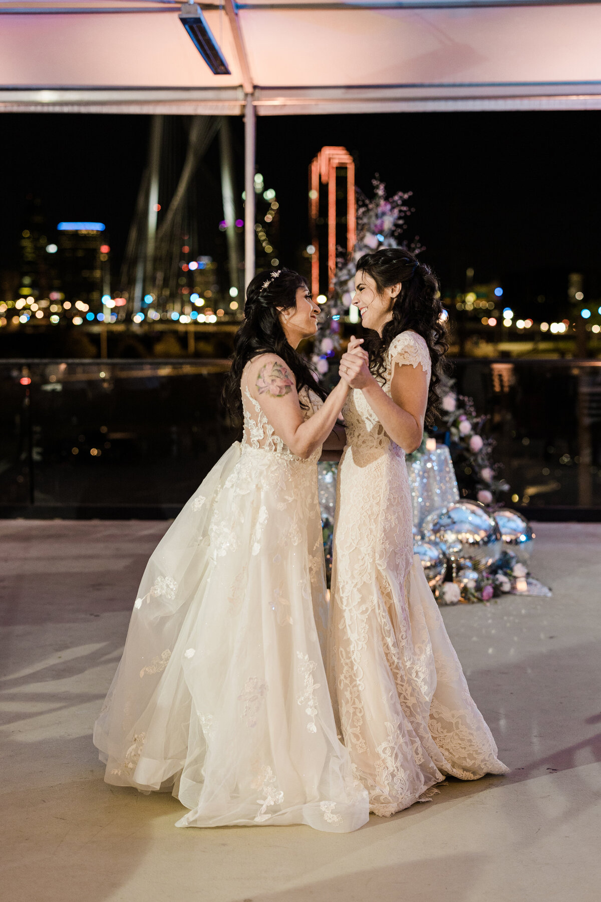 A candid shot of two brides sharing their first dance during their wedding reception in downtown Dallas, Texas. Both brides are smiling and looking joyfully into each other's eyes. Both brides are wearing intricate, white dresses, and the Dallas skyline can be seen lit up in the background.