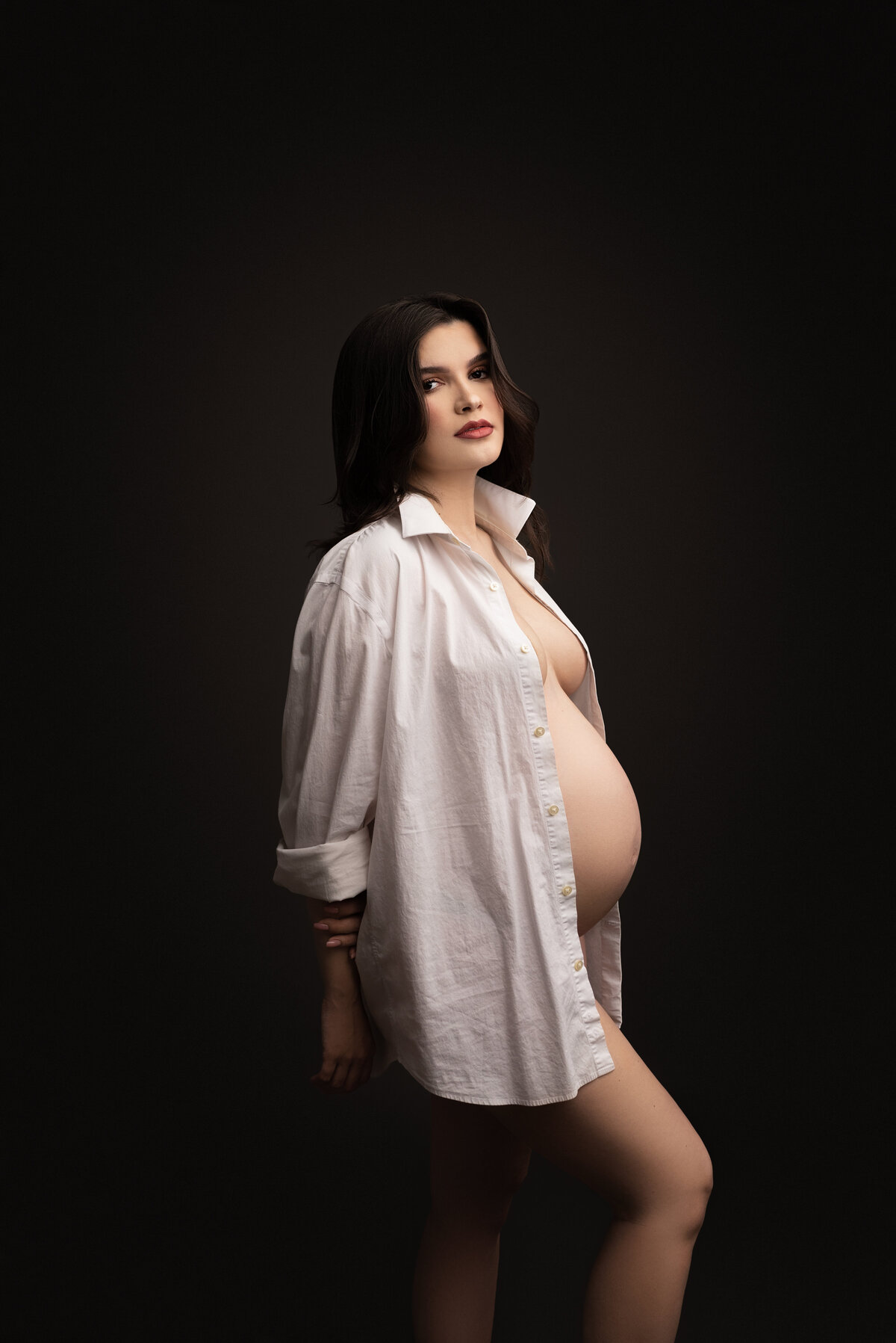 A woman with long dark hair and bold red lipstick stands angled to the camera for a fine art maternity photoshoot in Swedesboro, NJ. She confidently wears an unbuttoned dress shirt, revealing her beautiful baby bump. With an alluring glance over her shoulder towards the camera, she exudes both elegance and strength, creating a bold and memorable image captured by the best New Jersey maternity photographer, Katie Marshall.