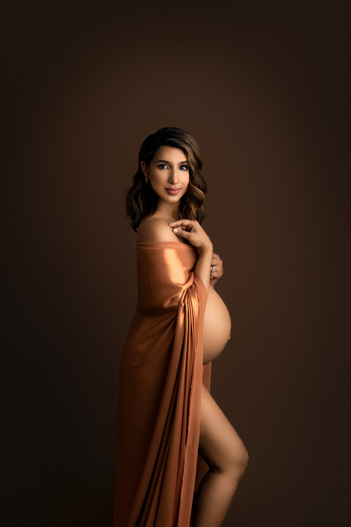 In this fine art photograph, Katie Marshall, the renowned Main Line maternity photographer, artistically captures the allure of an expectant mom. The radiant mother stands gracefully in a stunning copper-colored silk gown, her profile accentuated by the gentle drape of the fabric. Her flowing reddish-brown curls cascade elegantly over her shoulder, framing her serene expression as she gazes back over her shoulder towards the camera, her smile exuding warmth and anticipation.