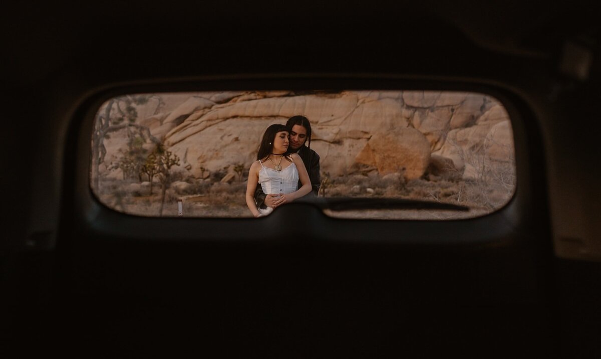 Couple embraces outside in the desert, photographed through the rear window of a car in Joshua Tree National Park.