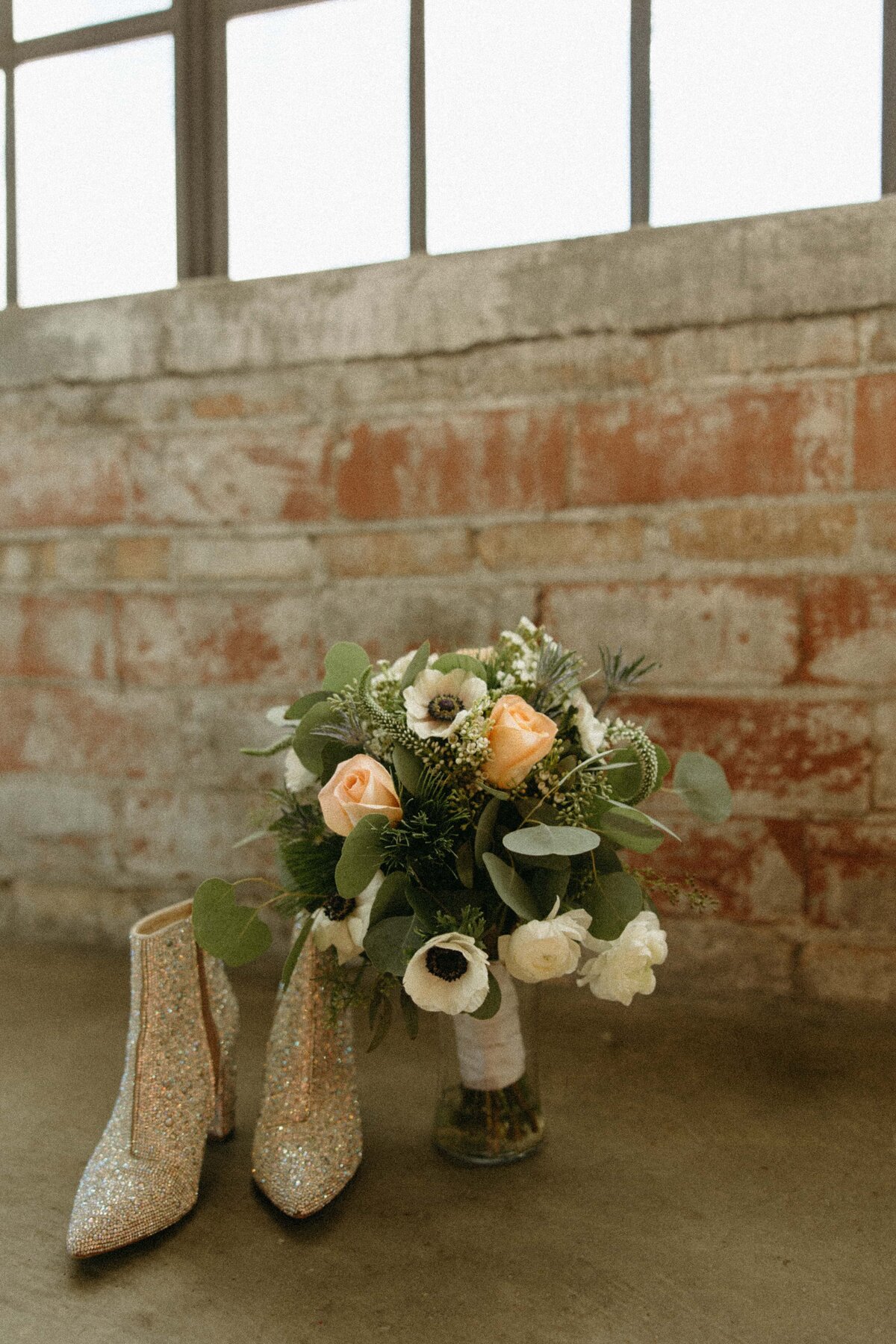 A bridal bouquet of white and soft pink flowers next to glittering high-heeled shoes on a concrete floor, with a brick wall and windows in the background at a Iowa wedding.