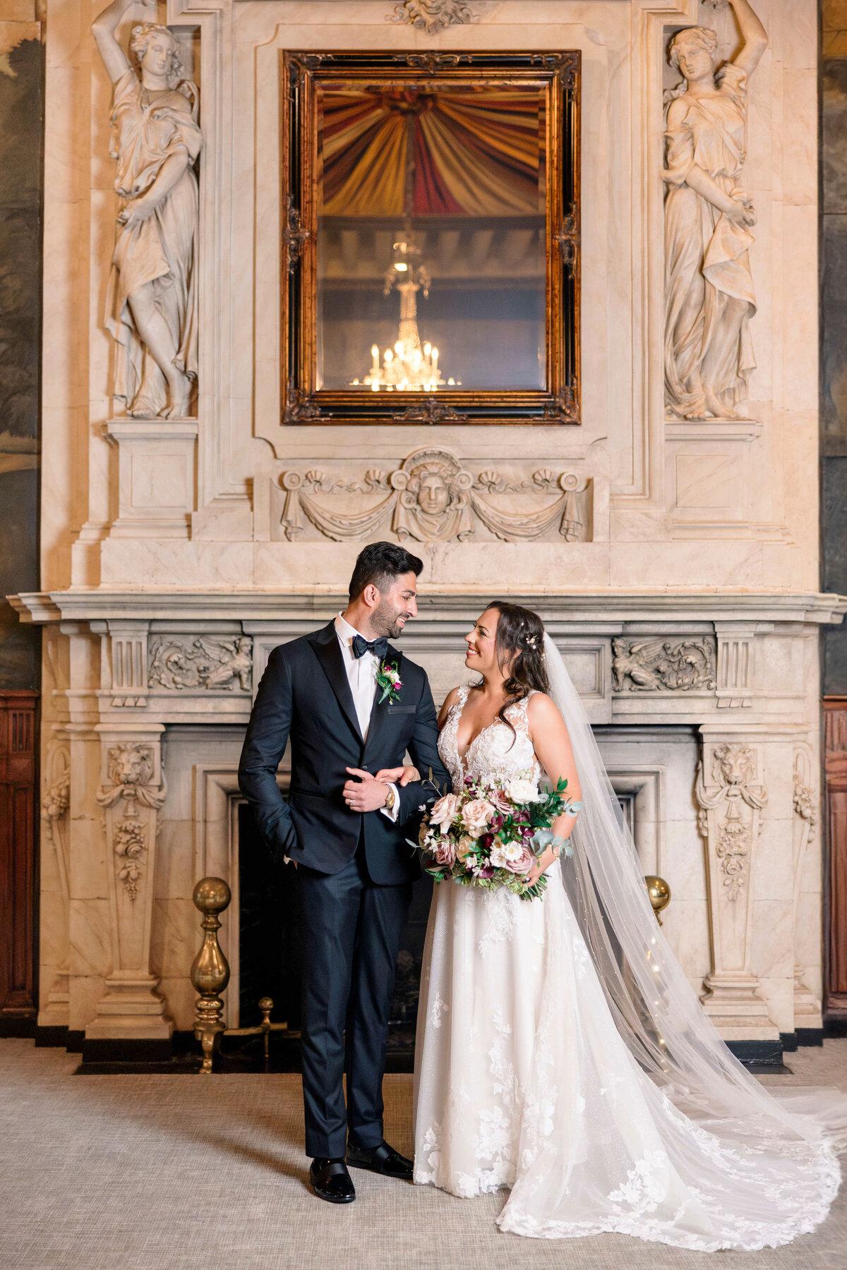 A bride and groom stand smiling at each other in front of an ornate fireplace. The bride holds a bouquet and wears a lace gown with a long veil. The groom is in a tuxedo. The setting is elegant, with statues and a chandelier reflected in a mirror above them.