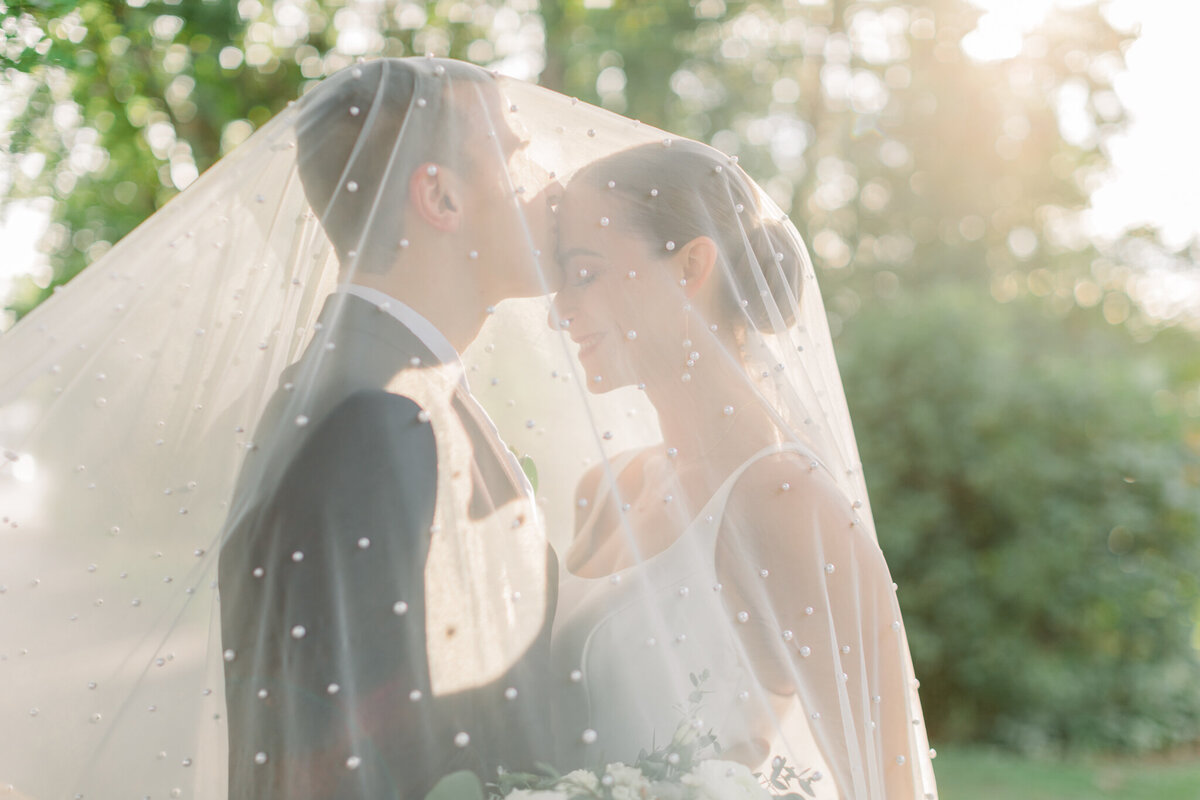 Bride and groom in soft, romantic sunset light under a pearl studded veil - groom is kissing the bride's forehead while she smiles
