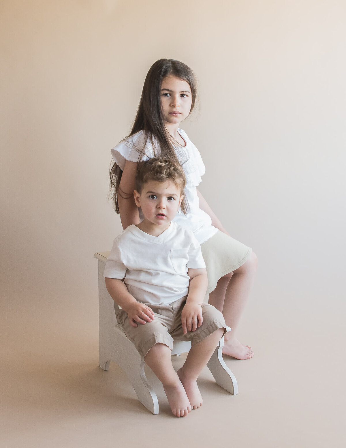 siblings wearing beige bottoms and white tops  with serious faces  with a tan background