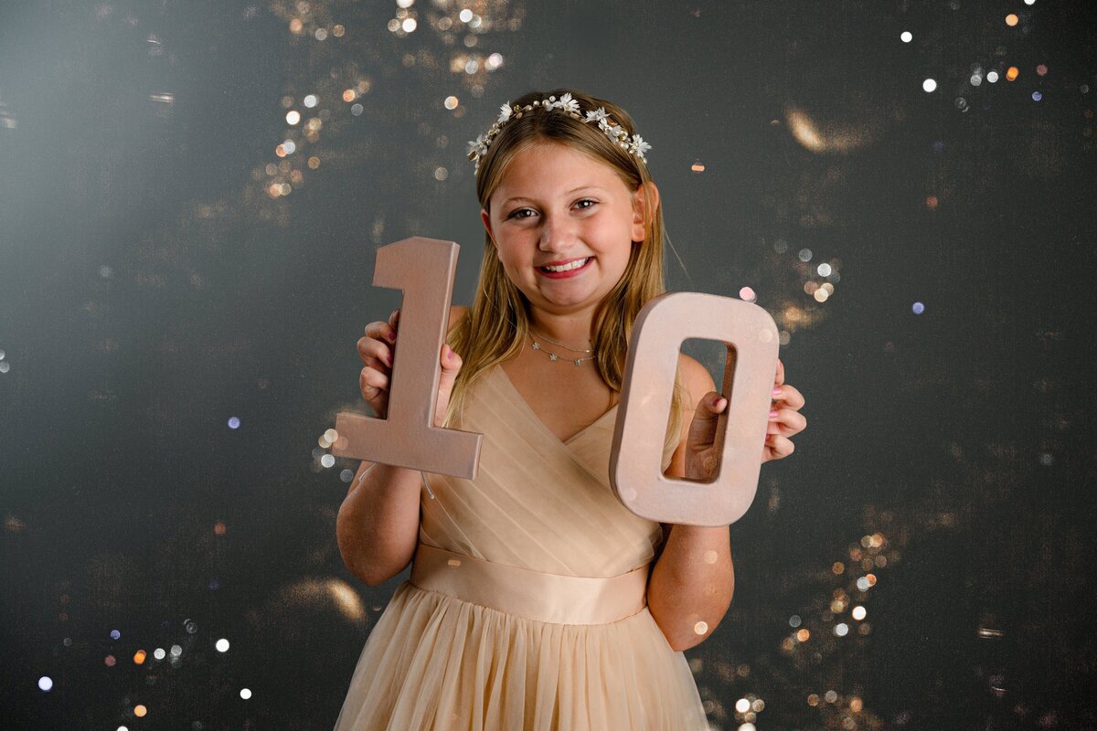 Little girl dressed in pink holding a one and zero for her 10th birthday Milestone session surrounded by glitter on a gray background