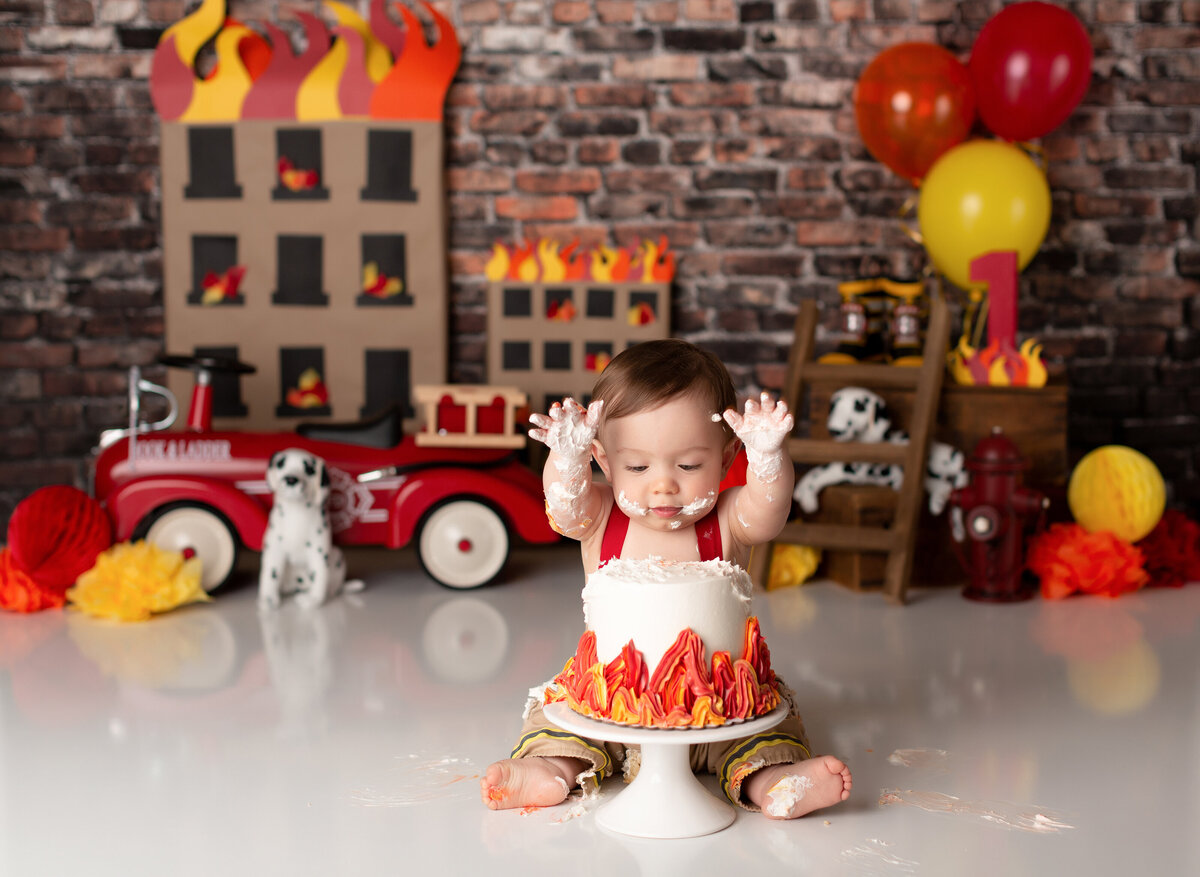 Firefighter themed cake smash in West Palm Beach and Boca Raton photography studio. Baby boy about to smash both hands in a white cake decorated with flames. The background has exposed brick wall, vintage toy firetruck, ladders, and Dalmatians.