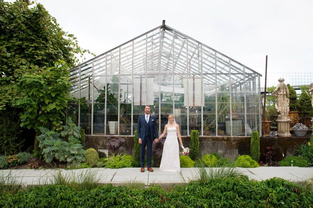 a bride and groom hold hands looking at the camera standing in front of the greenhouse at pomarius garden