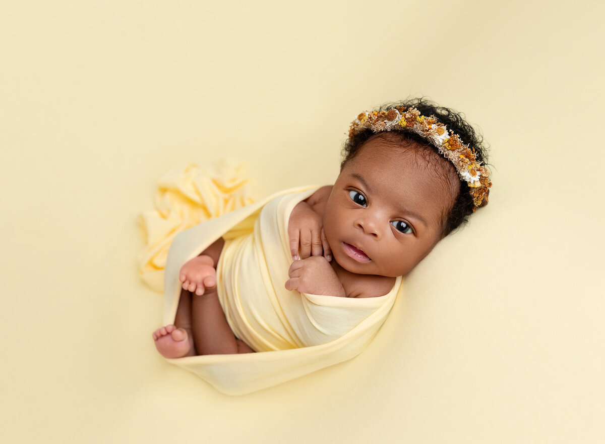 Baby girl is awake for a newborn photoshoot. She is wrapped in a yellow swaddle with her hands and toes peeking out. She is wearing a yellow floral crown. She is looking at the camera.