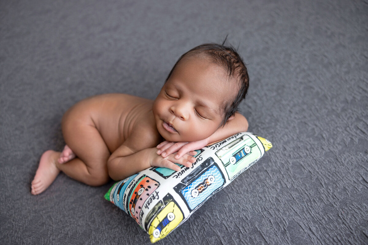 A sleeping baby lies peacefully on a small, colorful pillow with cartoon cars, perfectly captured by a Jacksonville newborn photographer. The baby has their arms gently resting under their head, set against a soft gray fabric backdrop.